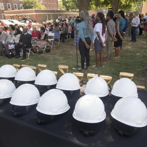 White hard hats on a table while a crowd gathers at the Paul Robeson Plaza groundbreaking